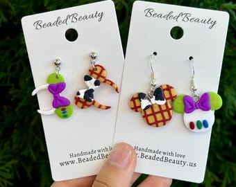Könnte beinhalten: Ein Paar handgefertigte Ohrringe aus Polymer Clay mit einem rot-gelb karierten Mickey Mouse-Ohren-Design mit einer schwarz-weißen Kuhmuster-Schleife und einem grün-weißen Mickey Mouse-Ohren-Design mit einer lila Schleife und bunten Punkten. Die Ohrringe befinden sich auf weißen Karten mit dem Text "Beaded Beauty" und Website-Informationen.