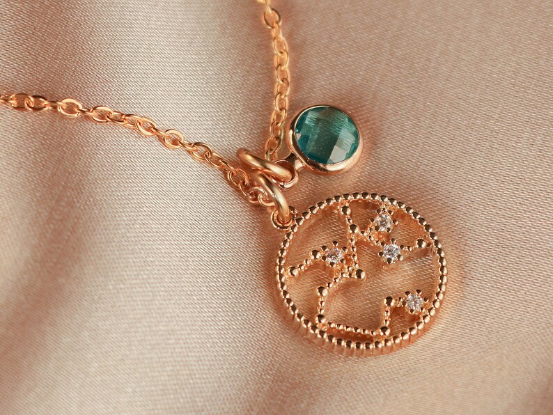 May include: A gold necklace with a round pendant featuring a constellation design with small clear stones. The pendant is attached to a small teal blue gemstone.