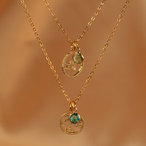 May include: Two gold necklaces with delicate chains and circular pendants. The top pendant features a green gemstone and small sparkling stones. The bottom pendant features a blue gemstone and sparkling stones.