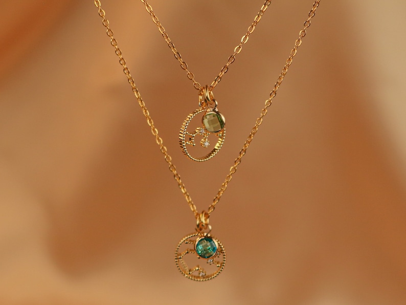 May include: Two gold necklaces with delicate chains and circular pendants. The top pendant features a green gemstone and small sparkling stones. The bottom pendant features a blue gemstone and sparkling stones.