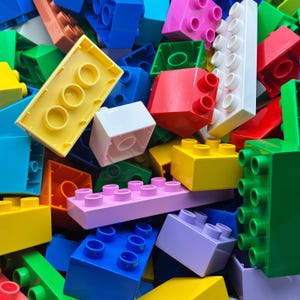 May include: A close-up of a collection of brightly coloured plastic building blocks. The blocks are in shades of yellow, red, green, blue, pink, and white. The blocks vary in size and shape, with studs on top for connecting.