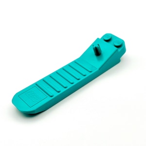 May include: A teal LEGO brick separator tool. The tool has a long, flat body with a series of parallel ridges and the LEGO logo. The end has a small, angled section with two studs and a central post.