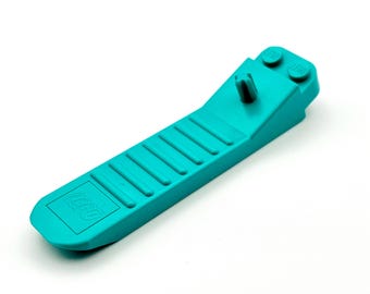 Peut inclure: Un séparateur de briques LEGO de couleur turquoise. L'outil a un corps long et plat avec une série de rainures parallèles et le logo LEGO. L'extrémité comporte une petite section inclinée avec deux tenons et un poteau central.
