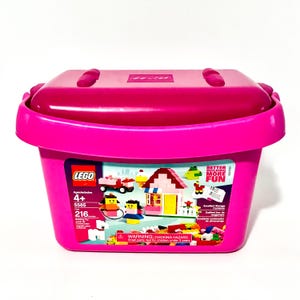 May include: A pink LEGO storage container with a matching lid. The container features a colorful illustration of a house, figures, and vehicles. The text on the container reads "LEGO", "Better Building More Fun", and "Warning: Choking Hazard".