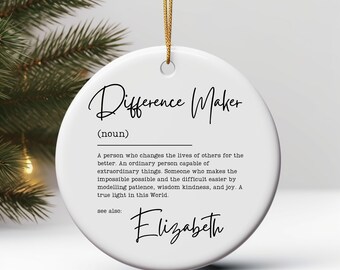 May include: White ceramic ornament with the text "Difference Maker" in a script font. The ornament includes a definition and the name "Elizabeth" in a script font. A gold string is attached for hanging.
