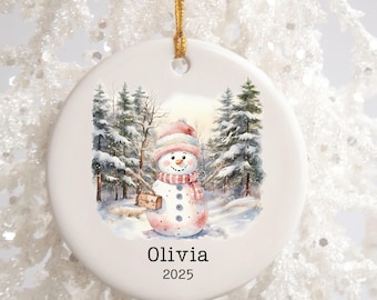 May include: A white ceramic Christmas ornament featuring a watercolor illustration of a snowman in a winter forest scene. The snowman wears a pink hat and scarf, and carries a small bag. The name "Olivia" and the year "2025" are printed below.