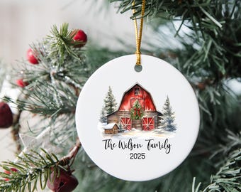 May include: A white ceramic Christmas ornament featuring a watercolor painting of a red barn with snow and trees. The text "The Wilson Family 2025" is written in black script. A gold string is attached for hanging.