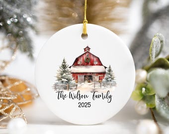 May include: A white ceramic Christmas ornament with a gold string. The ornament features a watercolor painting of a red barn in a snowy landscape, with the text "The Wilson Family 2025".