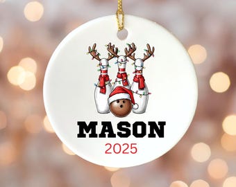 May include: A white ceramic Christmas ornament with a gold string. The ornament features three bowling pins decorated as reindeer with red scarves and Christmas lights. A bowling ball wears a Santa hat. The name "MASON" and the year "2025" are printed below.