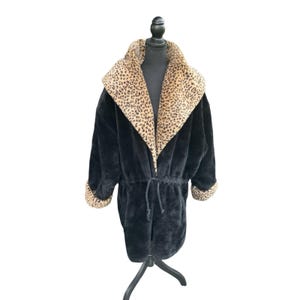 May include: A black faux fur coat featuring a wide shawl collar with a tan and black leopard print pattern. The coat includes long sleeves with matching leopard print cuffs and a drawstring waist with black cords for an adjustable fit.