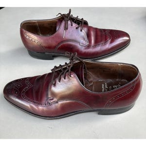 May include: A pair of burgundy leather wingtip oxford shoes featuring decorative brogue detailing on the toe cap and along the sides. The shoes have dark brown laces, a low heel, and a smooth polished finish with visible interior branding.