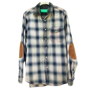 May include: A long-sleeve button-down shirt featuring a navy blue and off-white plaid pattern. The shirt includes a single chest pocket, a collar, and brown suede-like elbow patches on each sleeve. A green United Colors of Benetton label is visible.
