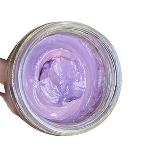 Peut inclure: Un pot en verre transparent rempli d'une crème épaisse, lisse et de couleur violet clair. La substance présente une consistance dense et opaque avec des tourbillons et des pics visibles en surface, contenue dans un récipient circulaire.