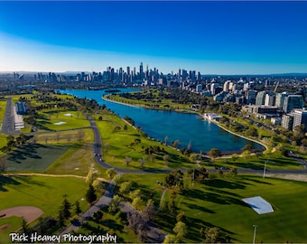 May include: An aerial view of a city skyline with a large lake in the foreground. The lake is surrounded by green grass and trees. The city skyline is in the background and is made up of tall buildings. The sky is blue and clear.