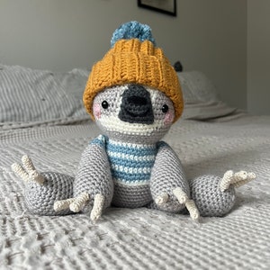 May include: A crocheted sloth wearing a yellow and blue hat with a pom-pom. The sloth is sitting on a gray knitted blanket.