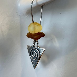May include: A single earring with a silver hook. The earring features a yellow bead, a brown, irregular-shaped bead, and a silver triangle pendant with a spiral design. The pendant has a textured surface.