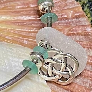 Könnte beinhalten: Ein silbernes Armband mit einem keltischen Knotenanhänger, Seeglasperlen und Silberperlen. Das Armband wird auf einer Muschel und einer weißen Oberfläche präsentiert. Die Seeglasperlen sind helltürkis.