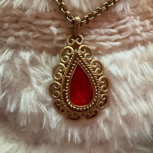 May include: A gold-toned metal pendant necklace featuring a teardrop-shaped red stone set within an ornate, filigree-style frame. The pendant is suspended from a matching gold-toned chain with textured, rounded links that connect to the top bail.