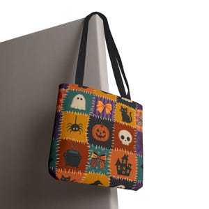 May include: A tote bag with black handles featuring a patchwork pattern of Halloween icons including a ghost, orange pumpkin, black cat, spider, skull, cauldron, haunted house, and bows on orange, purple, teal, and brown square backgrounds.