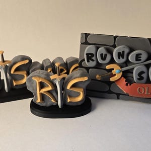 Pu&ograve; includere: Tre oggetti decorativi con le parole "RuneScape" e "Old School". Gli oggetti sono grigi con scritte dorate e includono spade. Uno &egrave; un pezzo lungo e orizzontale, un altro un pezzo circolare e il terzo un pezzo rettangolare.