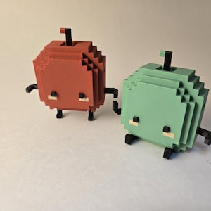 Puede incluir: Dos figuras pixeladas en forma de cubo, una roja y otra verde, con apéndices negros y pequeños detalles de color claro. Tienen un tallo en la parte superior y están diseñadas en un estilo retro de 8 bits, que recuerda a los personajes de videojuegos clásicos.