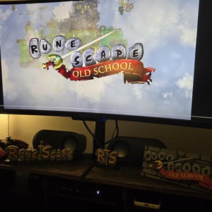 Pu&ograve; includere: Un monitor di computer mostra il logo del gioco "RuneScape Old School". Sotto il monitor, una mensola di legno espone altoparlanti, controller di gioco e oggetti decorativi a tema "RuneScape", tra cui un logo e una scena in miniatura.