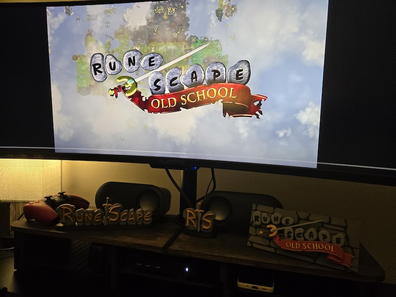 Pu&ograve; includere: Un monitor di computer mostra il logo del gioco "RuneScape Old School". Sotto il monitor, una mensola di legno espone altoparlanti, controller di gioco e oggetti decorativi a tema "RuneScape", tra cui un logo e una scena in miniatura.