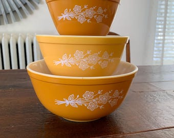 May include: Three vintage orange mixing bowls stacked on a wooden table. Each bowl has a white floral design. The bowls are different sizes, with the largest bowl at the bottom and the smallest bowl at the top.