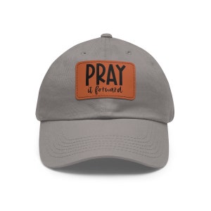 Może przedstawiać: Szara czapka z daszkiem z brązową skórzaną naszywką z napisem "Pray it forward" w czarnym kolorze.