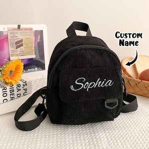 May include: A black corduroy mini backpack featuring a front pocket with the name Sophia embroidered in white script. The bag includes a top handle, adjustable black shoulder straps, and a small black D-ring attached to the front pocket flap.