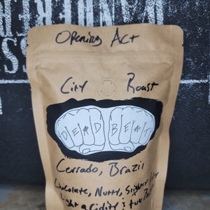 Może przedstawiać: Brązowa papierowa torba z nadrukiem słów "Opening Act" i "City Roast". Torba zawiera czarno-białą ilustrację dwóch pięści z napisem "Dead Beat". Torba zawiera również tekst "Cerrado, Brazylia", "Czekolada, Orzechowa, Lekko Lekka Kwasowość, 3 dwa"