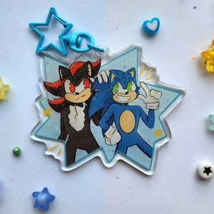 May include: A clear acrylic keychain featuring cartoon characters Sonic and Shadow. The keychain is in a starburst shape with a blue star-shaped clasp. Scattered around are small star and heart-shaped beads in various colors.