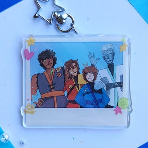 May include: A clear acrylic keychain featuring a cartoon illustration of five people in a photo booth. The photo booth has a white border and a pink heart in the corner. The people are all smiling and wearing different colored clothing. The keychain has a silver star charm attached to it.