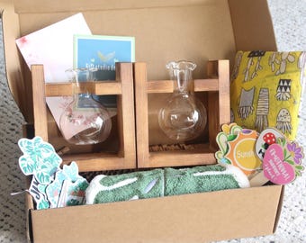 May include: A gift box containing two clear glass vases in wooden holders, a green towel, and decorative stickers. The box also includes a yellow fabric bag with a mushroom pattern and small cards. The box is made of cardboard.