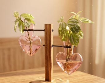 May include: A wooden plant stand with two heart-shaped glass vases. Each vase holds a small plant with green leaves and pink-tinted water. The stand is made of dark wood and sits on a wooden table.