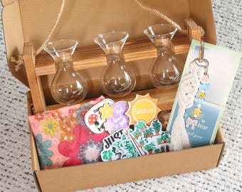 May include: A gift box with a wooden rack holding three small glass vases. The box contains stickers, a floral print fabric, and a macrame keychain with a card. The stickers have the words "Sunshine" and "Just".