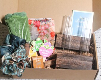 May include: A gift box filled with various items, including a green washcloth, floral print pouch, scrunchies, and a wooden vase with glass tubes. Decorative signs with text are also visible.