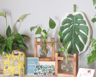 May include: A display of home decor items featuring plants and botanical themes. Includes small glass vases with plants in wooden stands, a yellow cactus-print pouch, and a green leaf-shaped wall hanging. Several cards with botanical designs are also present.