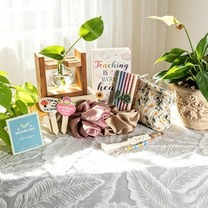 May include: A collection of items including a glass plant propagation bulb in a wooden stand, a notebook with the text Teaching is work heart, a set of pens, two satin scrunchies, a floral embroidered pouch, and a macrame keychain with beads.