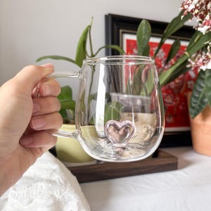 Puede incluir: Una taza de vidrio transparente con un detalle en forma de corazón en la base. La taza tiene un asa y un cuerpo redondeado. El corazón es de color rosa claro. La taza se sostiene en una mano.