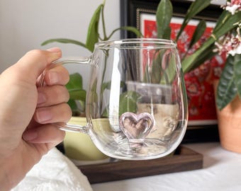 May include: A clear glass mug with a heart-shaped detail at the base. The mug has a handle and a rounded body. The heart is a soft pink color. The mug is held in a hand.