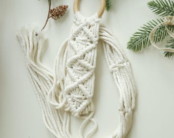 May include: A white macrame plant hanger with a wooden ring at the top. The hanger is made of thick cotton cord and has a diamond pattern.