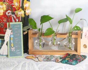 May include: A wooden plant stand with three glass vases, each holding a green plant. The stand is made of light brown wood and has a rope detail. A decorative macrame keychain and a small sign with text are on the left.