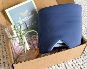 May include: A gift box containing a navy blue sleep mask, a clear glass with a wooden base, and a card that says "Gift studio decor". The glass is tied with a green ribbon. The box is filled with pink paper shreds.