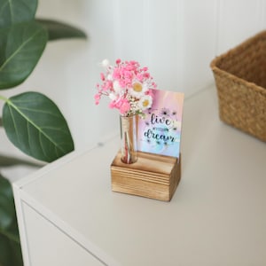 Puede incluir: Un pequeño bloque de madera sostiene un vial de vidrio con flores rosas y blancas. Una tarjeta con las palabras "live your dream" está al lado de las flores. El arreglo está sobre una superficie blanca.