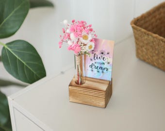 May include: A small wooden block holds a glass vial with pink and white flowers. A card with the words "live your dream" is next to the flowers. The arrangement sits on a white surface.