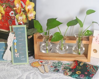 May include: A wooden plant stand holding three glass vases with green plants. A macrame keychain with colorful beads and a tassel hangs near a card with text. Stickers and a patterned pouch are also visible.