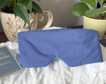 May include: A blue eye mask with a white interior rests on a white lace surface. The mask is rectangular with curved edges to fit around the eyes. The background includes potted plants and a decorative card.