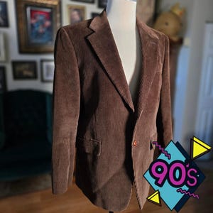 May include: A brown corduroy blazer displayed on a mannequin. The blazer has a notched lapel, a single-button closure, and a chest pocket. The image has a 90's graphic in the bottom right corner. The blazer is a medium brown color.