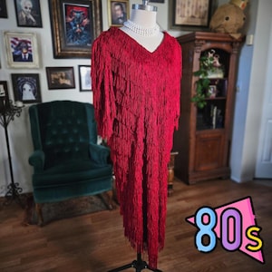 May include: A vibrant red fringed dress with a V-neck, displayed on a mannequin. The dress has a layered fringe design and is styled with a pearl necklace. The image includes a colorful "80s" graphic in the corner, suggesting a vintage theme.